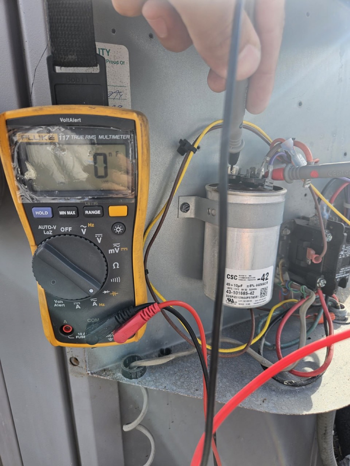 A Fluke 117 multimeter reading zero microfarads across a run capacitor labelled 45+10 uF, mounted in an open condenser electrical compartment